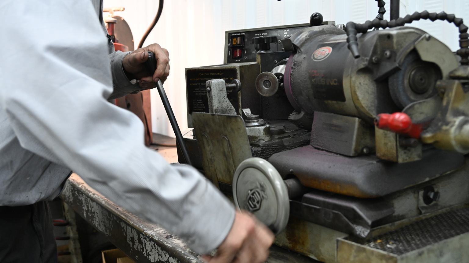 A PDM machinist operating a valve refacing machine, one hand on the feed lever and one on the handwheel.